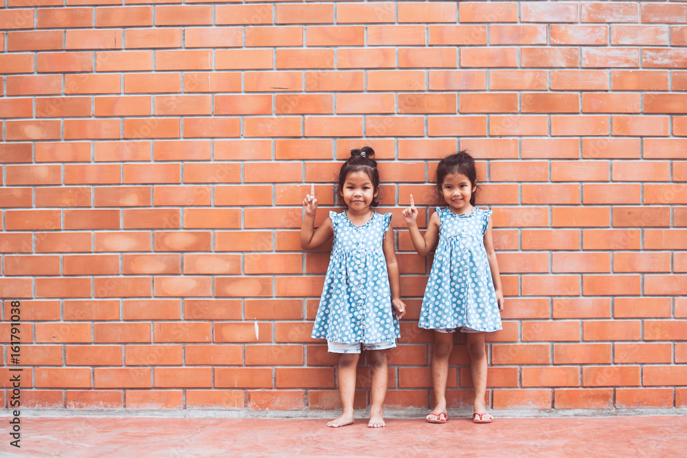 Portrait of two asian kid girls pointing up on brick wall background in vintage color tone Portrait of two asian kid girls pointing up on brick wall background in vintage color tone