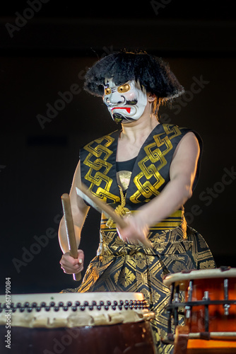 headache concept. devil drummer plays drumroll on the nerves. drummer girl in white demon mask with drumsticks, studio shot on a dark background. photo