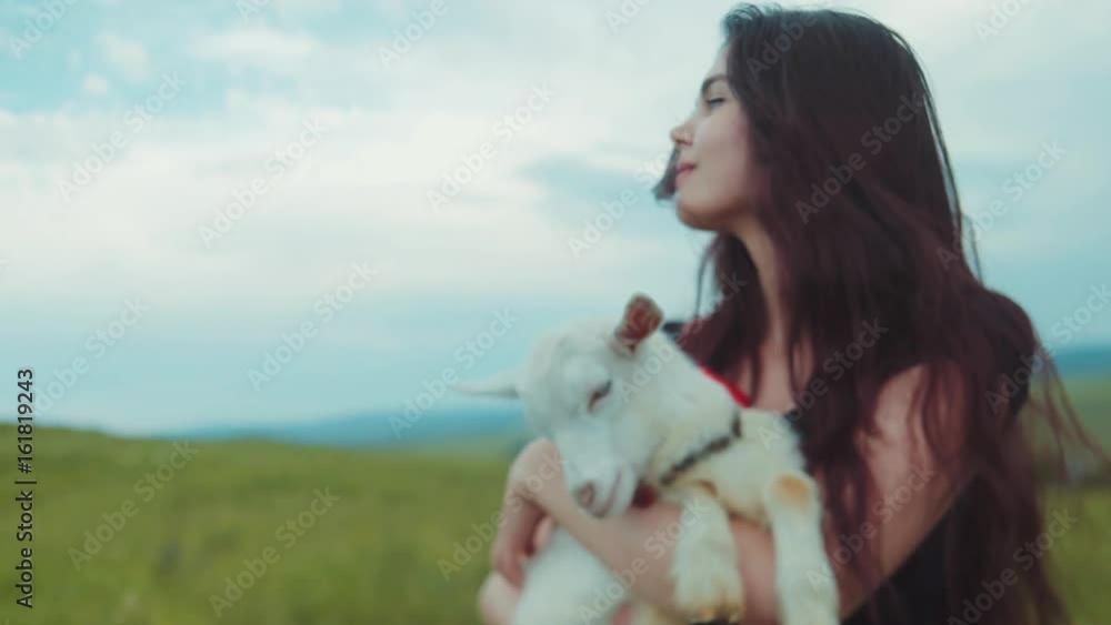 Beautiful long-haired brunette woman holds on her hands white goat kid, kisses it, smiles, pets and strokes it. Loving nature, green peace. Playful mood, being happy. Childhood memories. Beautiful long-haired brunette woman holds on her hands white goat kid, kisses it, smiles, pets and strokes it. Loving nature, green peace. Playful mood, being happy. Childhood memories.