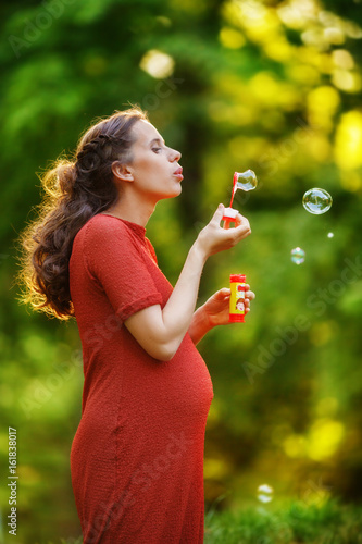 Pregnant woman in a red knitted dress blows soap bubbles  standing on the edge of the forest at sunset