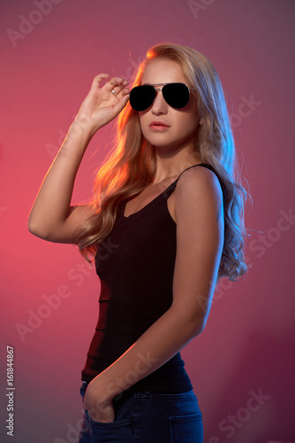 Portrait of young blonde woman wearing black top and sunglasses posing against red background