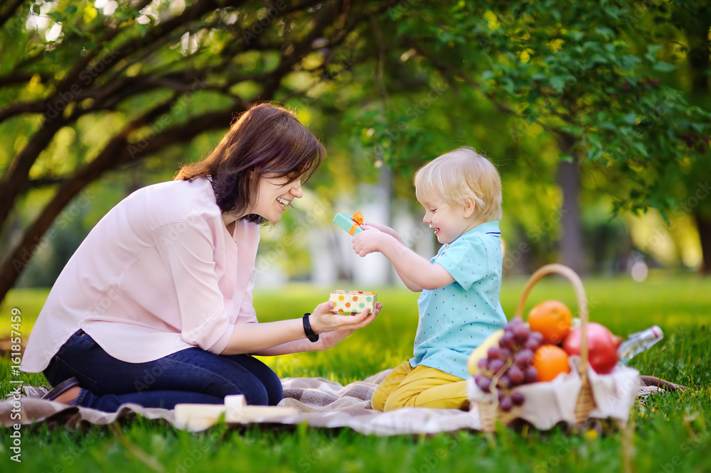 Cute little boy with his young mother opening nicely wrapped gift during picnic in sunny park Cute little boy with his young mother opening nicely wrapped gift during picnic in sunny park