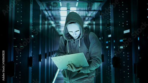 Schilderij op canvas Mid Shot of a Masked Hacker in a Hoodie Standing in the Middle of Data Center full of Rack Servers and Hacking it with His Laptop