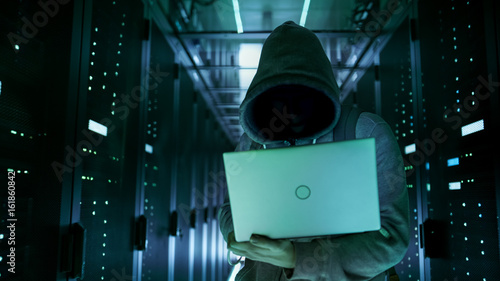 Close-up Shot of a Hacker with Hidden Face in a Hoodie Standing in the Middle of Data Center full of Rack Servers and Hacking it with His Laptop. photo