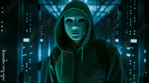 Obraz na plátně Masked Hacker in a Hoodie Standing in Corporate Data Center with Rows of Working Rack Servers