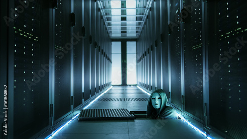 Fototapeta Naklejka Na Ścianę i Meble -  Hooded Hacker in a Mask Watching From Under Floor Hatch in Data Center with Rows of Rack Servers.