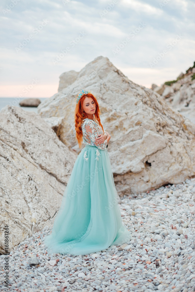 Young beautiful red-haired woman in a luxurious dress stands on a rocky shore of the Adriatic Sea among large stones