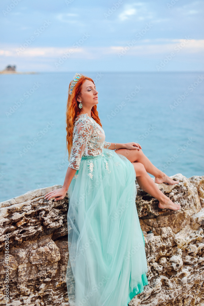 Young beautiful red-haired woman in mint dress sits on a large stone on the shore of the Adriatic Sea
