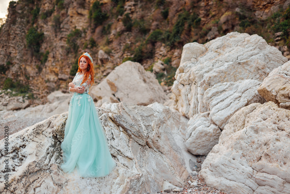 Young beautiful red-haired woman in a luxurious dress stands on a rocky shore of the Adriatic Sea among large stones