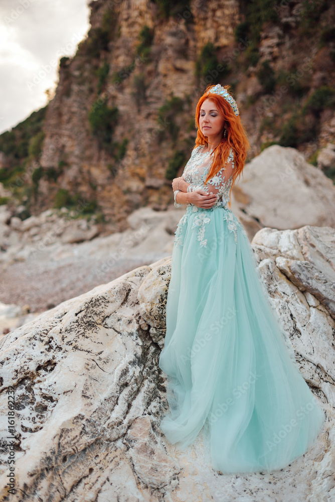 Young beautiful red-haired woman in a luxurious dress stands on a rocky shore of the Adriatic Sea among large stones Young beautiful red-haired woman in a luxurious dress stands on a rocky shore of the Adriatic Sea among large stones