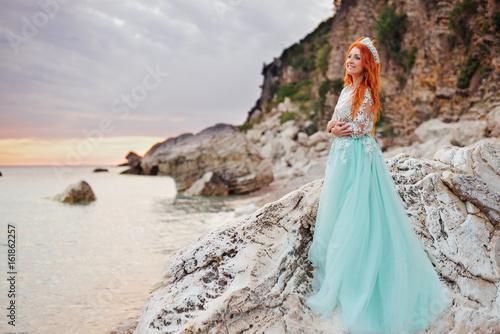 Young beautiful red-haired woman in a luxurious dress standing on a rocky shore of the Adriatic Sea