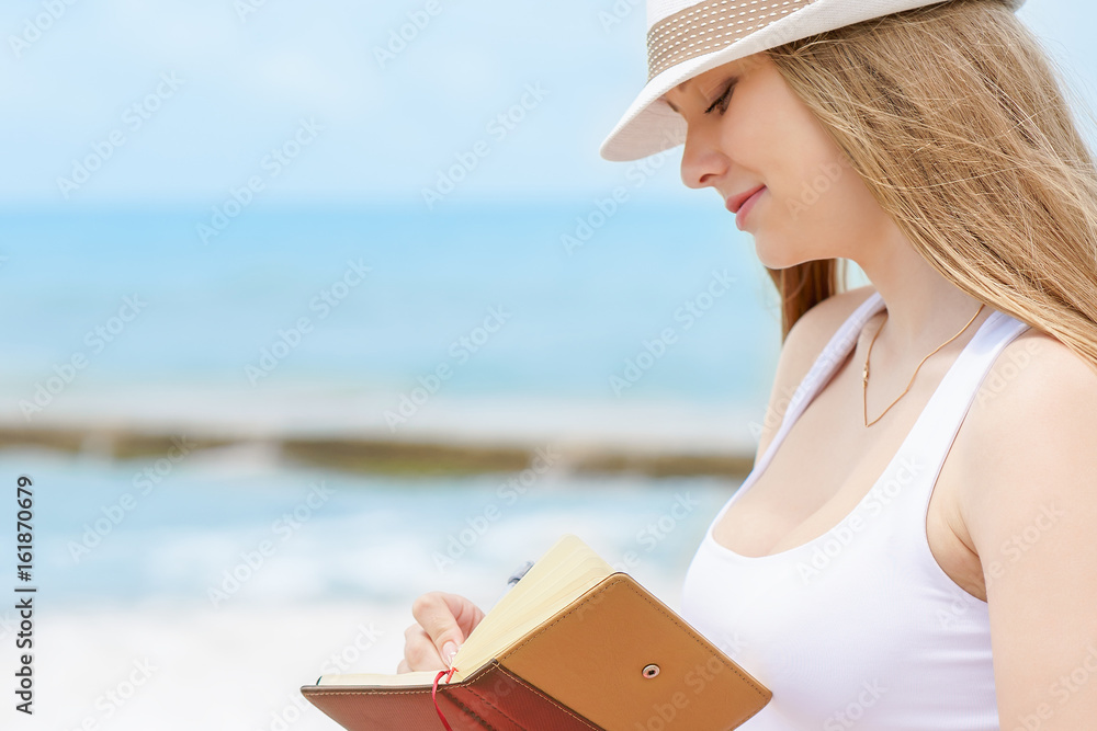 Young attractive girl with kind smile is writing some idea or letter in her note book by pen on background of blue sea
