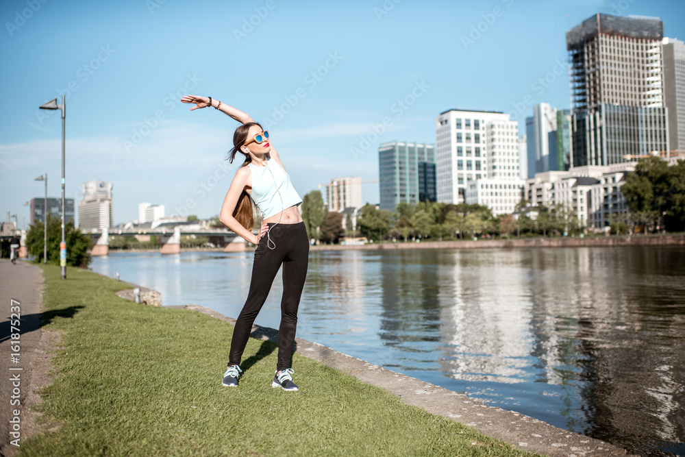 Fototapeta premium Young woman having a morning exercise stretching near the river with great view on the modern Frankfurt city