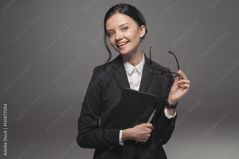 brunette businesswoman in black suit holding diary and smiling at camera, isolated on grey brunette businesswoman in black suit holding diary and smiling at camera, isolated on grey