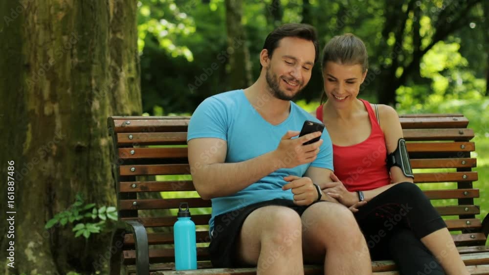 Happy young couple sitting on a bench after morning run and using smart phone. Man showing woman video, photos. Close up Happy young couple sitting on a bench after morning run and using smart phone. Man showing woman video, photos. Close up