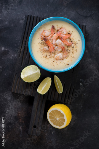 Black wooden serving board with a bowl of potato and shrimps cream-soup, high angle view on a dark scratched metal background