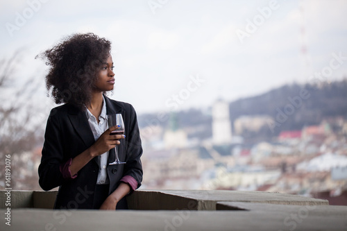 Attractive young black woman standing outside withthe glass of wine in her hands. Woman wearing suit. Great city view on the background. photo