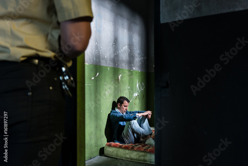 Young male prisoner sitting alone in an obsolete prison cell guarded by a police officer