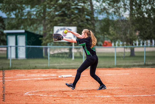 A teenage girl in fast ball pitching stance on the pitchers mound in black and green uniform