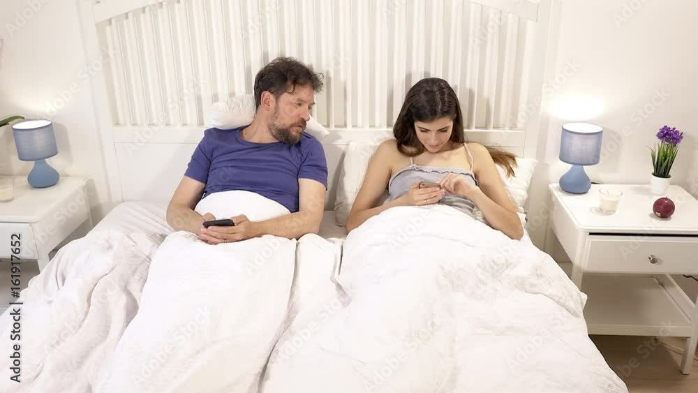 Jealous husband watching in bed wife writing text message with cell phone wide shot Jealous husband watching in bed wife writing text message with cell phone wide shot
