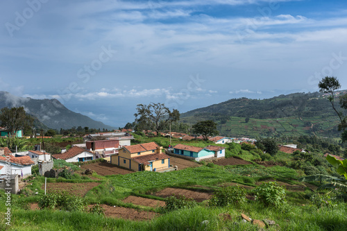 Nilgiri Hills, India - October 26, 2013: Thalaikunha hamlet with small farms and green and brown vegetable plots under blue sky. Highland scenery with mountains, dark green forests, colorful houses