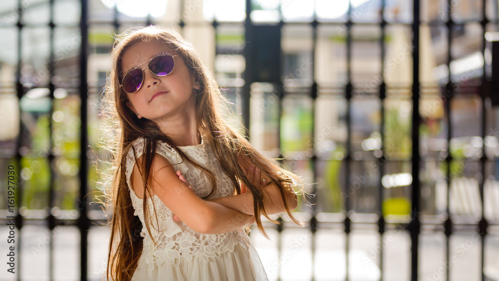 Portrait of a little girl in a white dress in sunglasses near the trellis fence. Close-up.