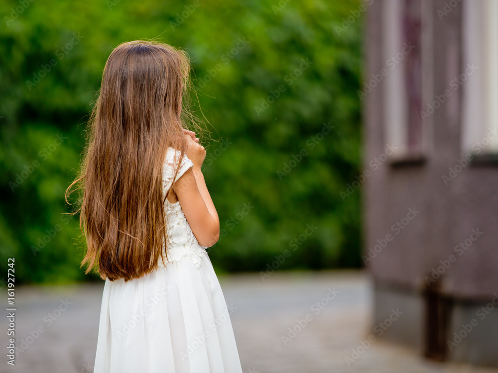 Naklejka premium Cute girl in white dress is standing with her back on a green fence background.