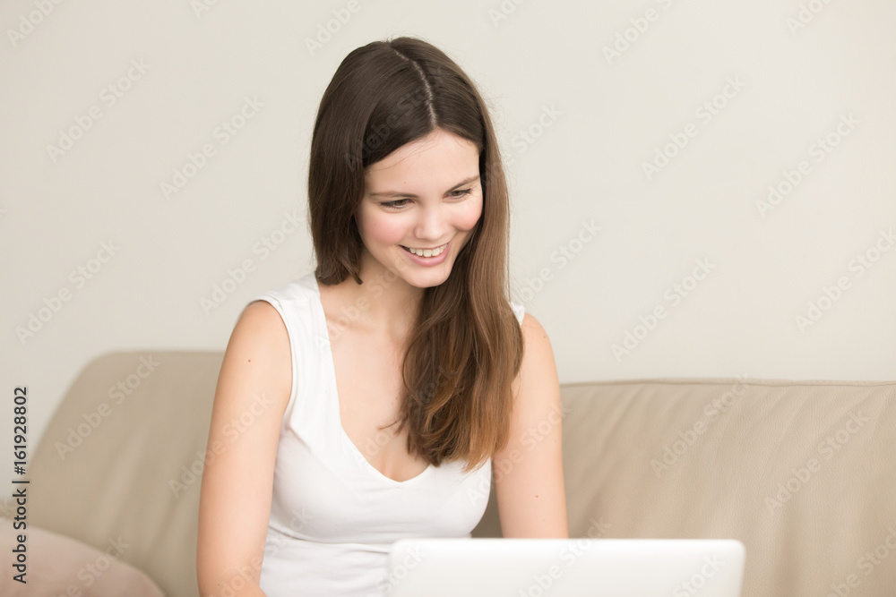 Smiling young woman sitting on sofa in front of laptop. Cheerful teen girl communicates with friend via skype, Internet, watching funny movie on computer. Female freelancer satisfied with work result Smiling young woman sitting on sofa in front of laptop. Cheerful teen girl communicates with friend via skype, Internet, watching funny movie on computer. Female freelancer satisfied with work result
