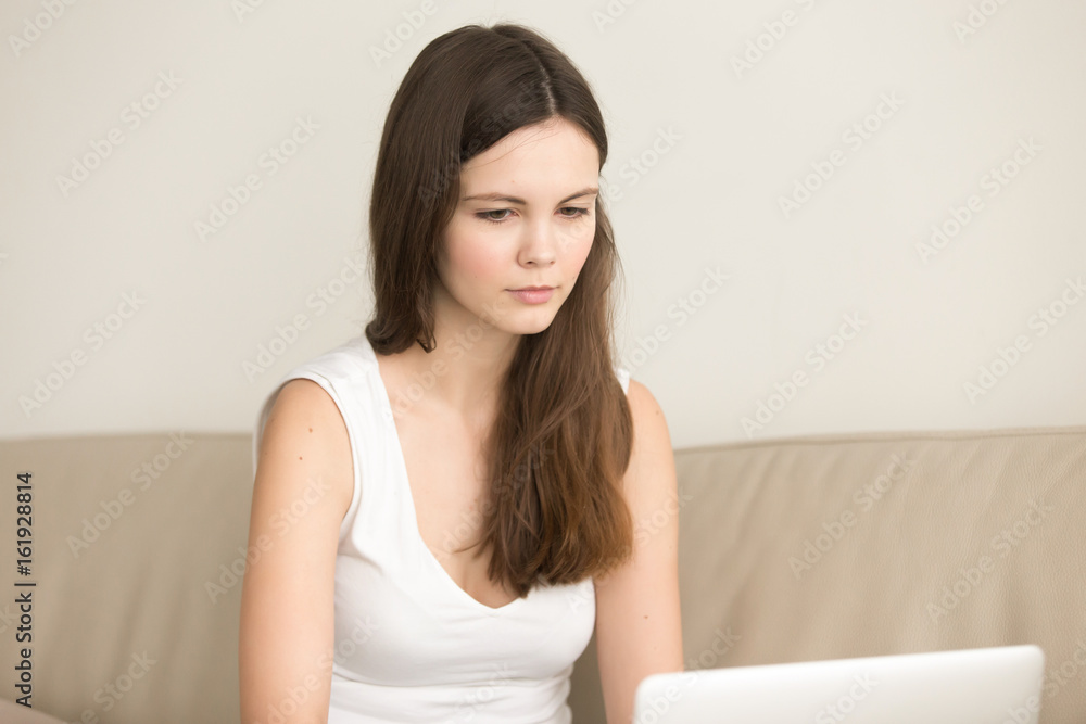 Beautiful young woman in casual clothing sitting on coach and looking on laptop screen. Cute calm lady working on computer at home, carefully studying information in Internet, selecting goods online Beautiful young woman in casual clothing sitting on coach and looking on laptop screen. Cute calm lady working on computer at home, carefully studying information in Internet, selecting goods online