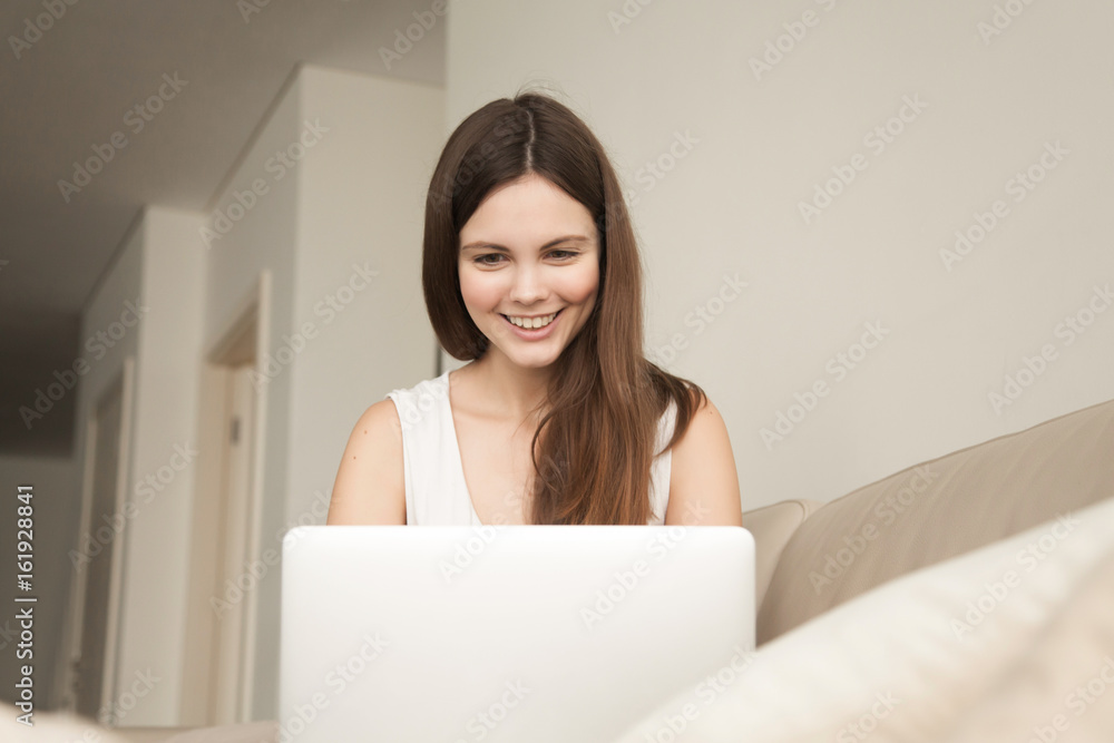 Beautiful young woman works on laptop on sofa at home. Smiling teen girl entertaining on computer, playing online game, communicating in Internet with friends. Happy lady using online banking service Beautiful young woman works on laptop on sofa at home. Smiling teen girl entertaining on computer, playing online game, communicating in Internet with friends. Happy lady using online banking service