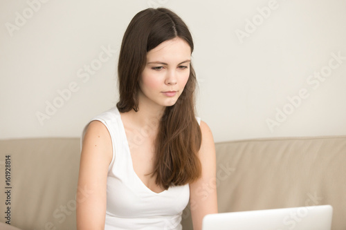 Beautiful young woman in casual clothing sitting on coach and looking on laptop screen. Cute calm lady working on computer at home, carefully studying information in Internet, selecting goods online
