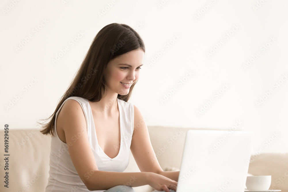 Attractive woman in casual clothes typing on laptop while sitting on sofa in bright living room. Smiling female entrepreneur or freelancer works distantly, communicates with colleague or client online Attractive woman in casual clothes typing on laptop while sitting on sofa in bright living room. Smiling female entrepreneur or freelancer works distantly, communicates with colleague or client online