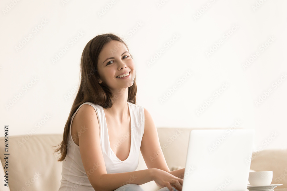 Portrait of happy smiling young woman sitting on sofa and using laptop. Lady surfing Internet, communicates in social network, satisfied with online shopping. Female teen enjoying work on computer Portrait of happy smiling young woman sitting on sofa and using laptop. Lady surfing Internet, communicates in social network, satisfied with online shopping. Female teen enjoying work on computer