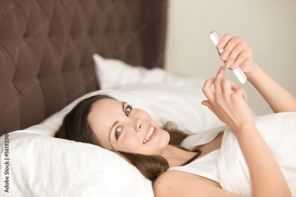 Portrait of young woman lying in bed using cellphone and looking in camera. Attractive female messaging with smartphone after waking up in morning at home, enjoying mobile app, reads news in network Portrait of young woman lying in bed using cellphone and looking in camera. Attractive female messaging with smartphone after waking up in morning at home, enjoying mobile app, reads news in network