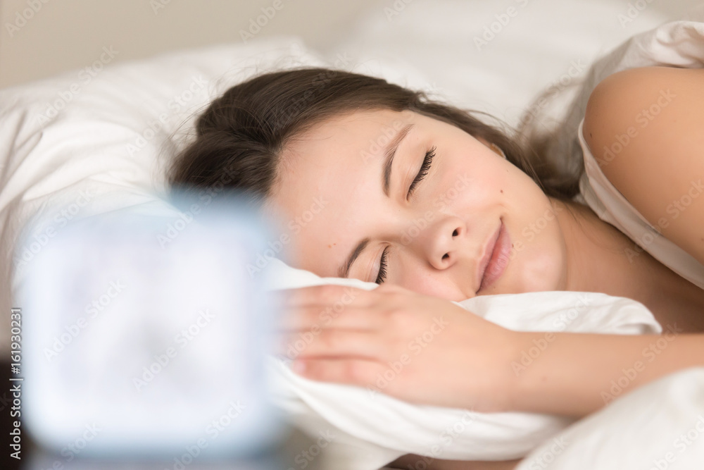Close up portrait of beautiful young woman sleeping in bad with blurred alarm clock on front plan. Adorable lady resting with pleasure at home bedroom, relaxing before going at work in the morning Close up portrait of beautiful young woman sleeping in bad with blurred alarm clock on front plan. Adorable lady resting with pleasure at home bedroom, relaxing before going at work in the morning