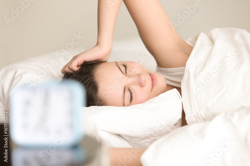 Young cute woman wakes up at morning in bed after alarm clock signal. Pretty lady feeling positive after good night sleep, awakening on comfortable bed with white linen. Easy morning wakeup concept