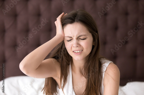 Young woman suffering from headache or migraine after wake up in morning.  Stressed lady in bed with painful face expression feels terrible hangover,  sharp pain in head, unpleasant weakness, dizziness Stock Photo