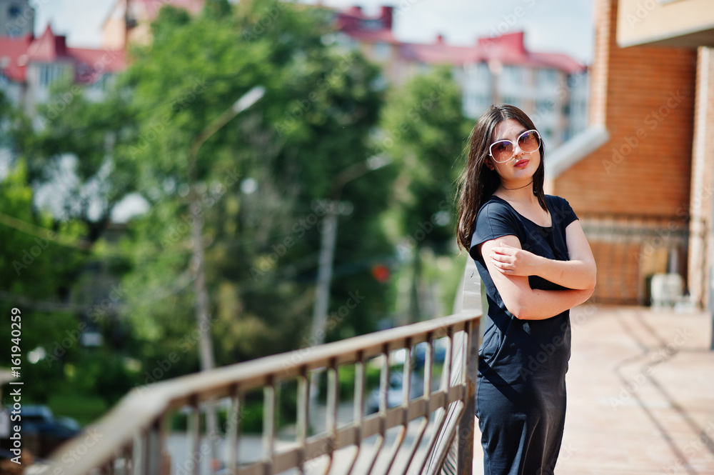 Brunette girl at black dress, sunglasses posing at street of city.