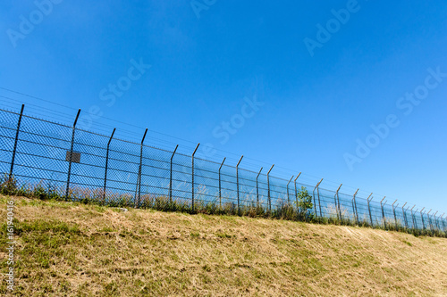 A border made of a double wire fence with barbed wire on top of a bank under a blazing sun and a summer blue sky.