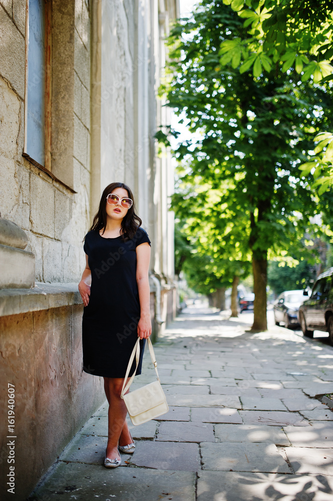 Brunette girl at black dress on sunglasses with handbag at hand posing at street of city.
