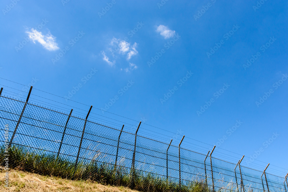 A border made of a double wire fence with barbed wire on top of a bank under a blazing sun and a summer blue sky. A border made of a double wire fence with barbed wire on top of a bank under a blazing sun and a summer blue sky.