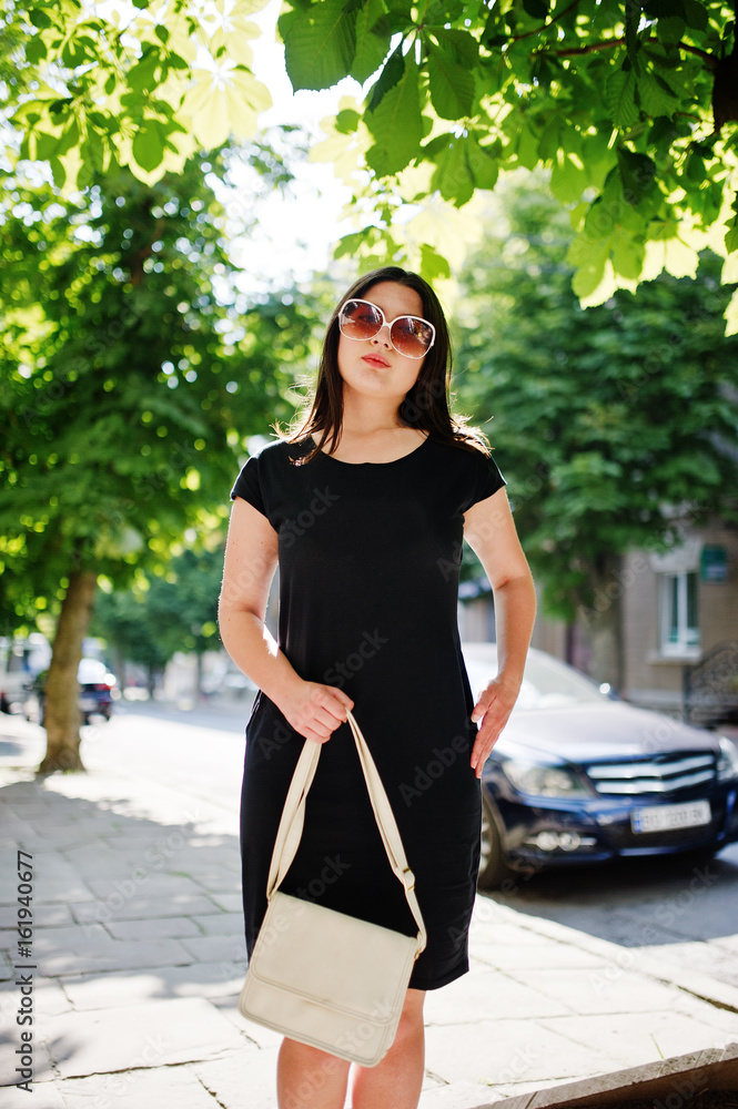 Brunette business girl at black dress on sunglasses with handbag at hand posing at street of city, background car on road. Brunette business girl at black dress on sunglasses with handbag at hand posing at street of city, background car on road.