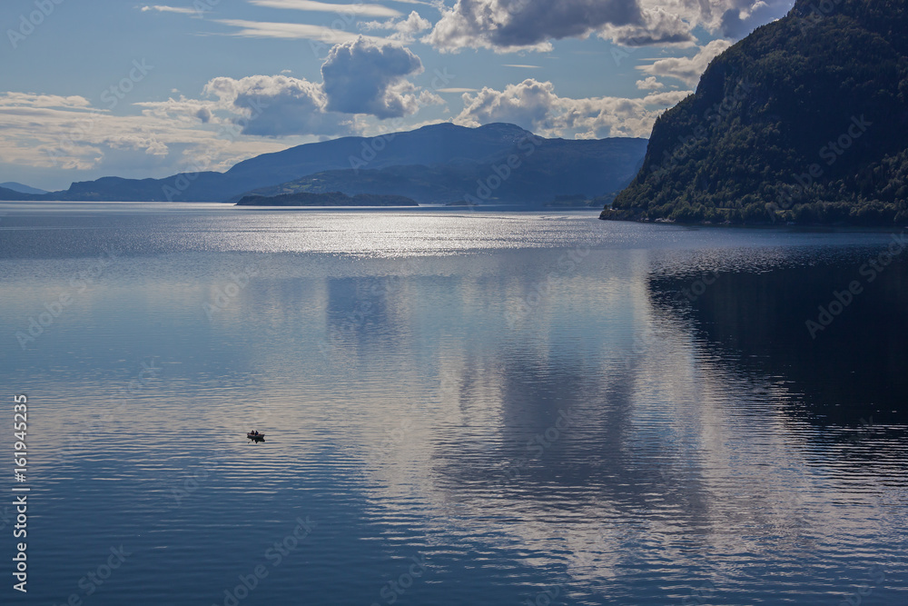 Small boat on the calm water mirror surface of the fjord on the beautiful majestic mountains background at evening time, Norway