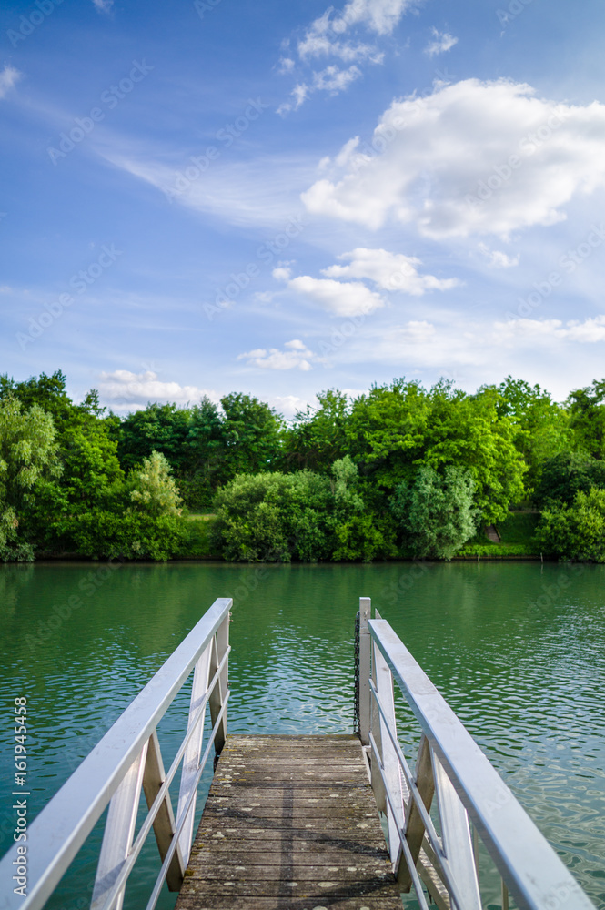 A wooden pontoon with metallic handrail over a river with trees and vegetation under a blue sky with white clouds.