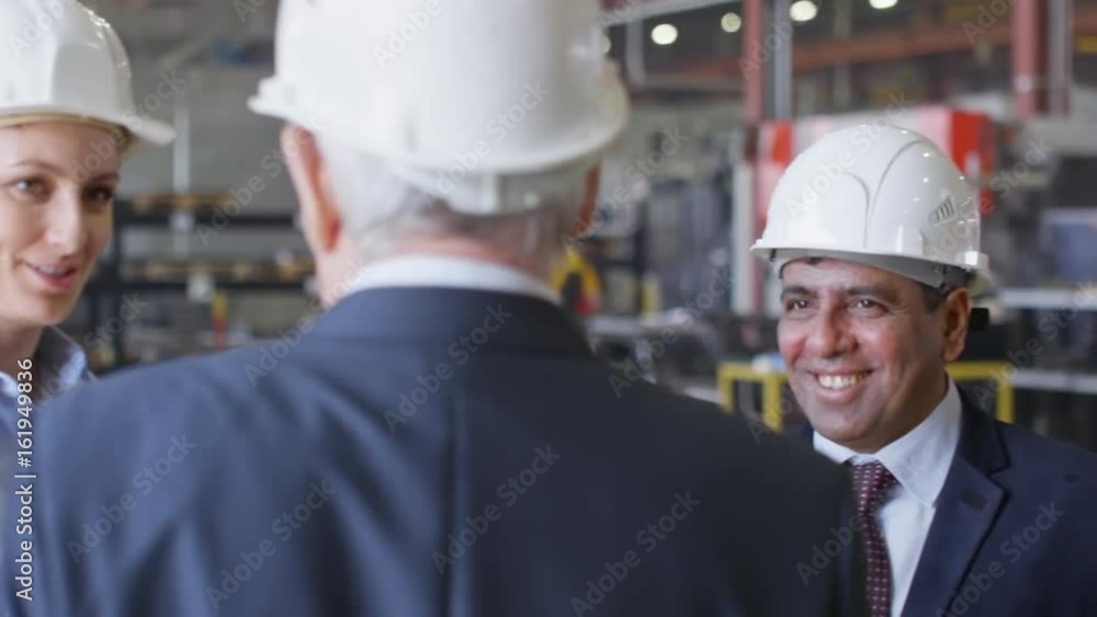 Tracking of multi-ethnic team of industrial engineers in formal wear and hardhats standing inside of industrial factory and discussing something Tracking of multi-ethnic team of industrial engineers in formal wear and hardhats standing inside of industrial factory and discussing something