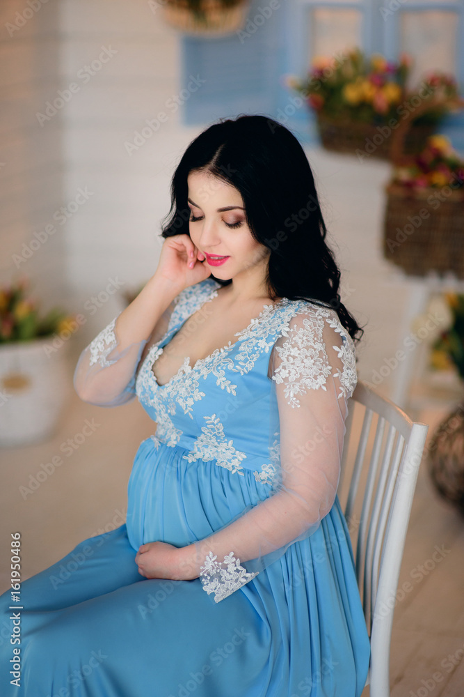 Gorgeous pregnant brunette in blue dress sits on white chair Gorgeous pregnant brunette in blue dress sits on white chair
