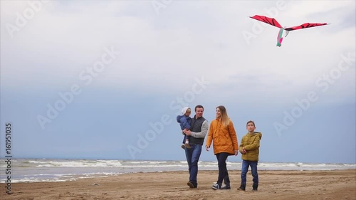 A young family walks along the beach next to the sea in cold weather, the son is holding a kite in his hands, the father carries a child, the mother is happy with a good weekend