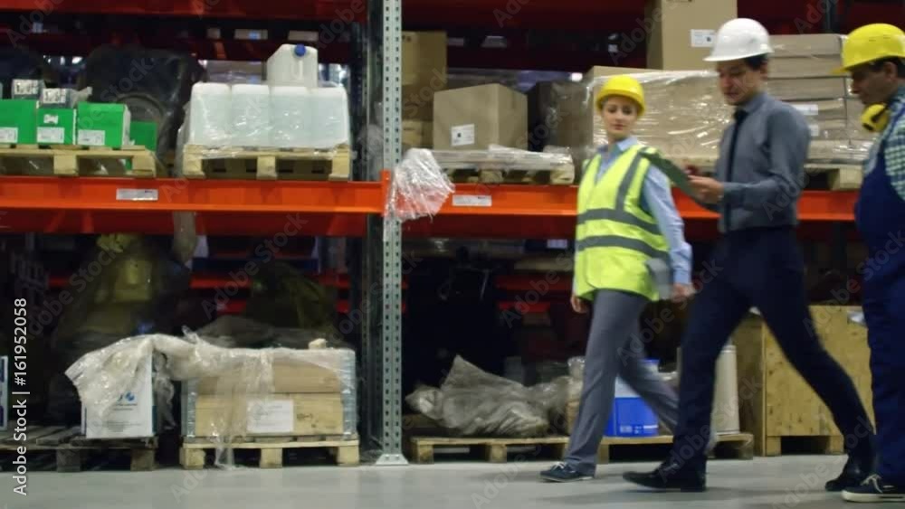 Side view of factory supervisor walking with two colleagues along shelves in warehouse in slow motion Side view of factory supervisor walking with two colleagues along shelves in warehouse in slow motion