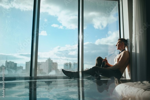 A man with a naked torso and a bottle of champagne sitting on the window