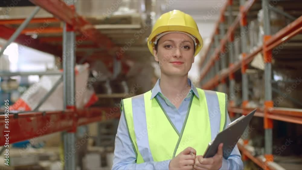Female engineer in visible uniform and hardhat walking in slow motion along shelves in warehouse and looking at camera Female engineer in visible uniform and hardhat walking in slow motion along shelves in warehouse and looking at camera
